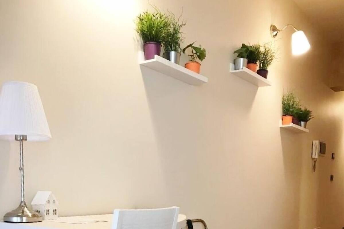 A room with a white table, a white chair, a small white house decoration, and a lamp. There are two white shelves with potted plants on the wall.