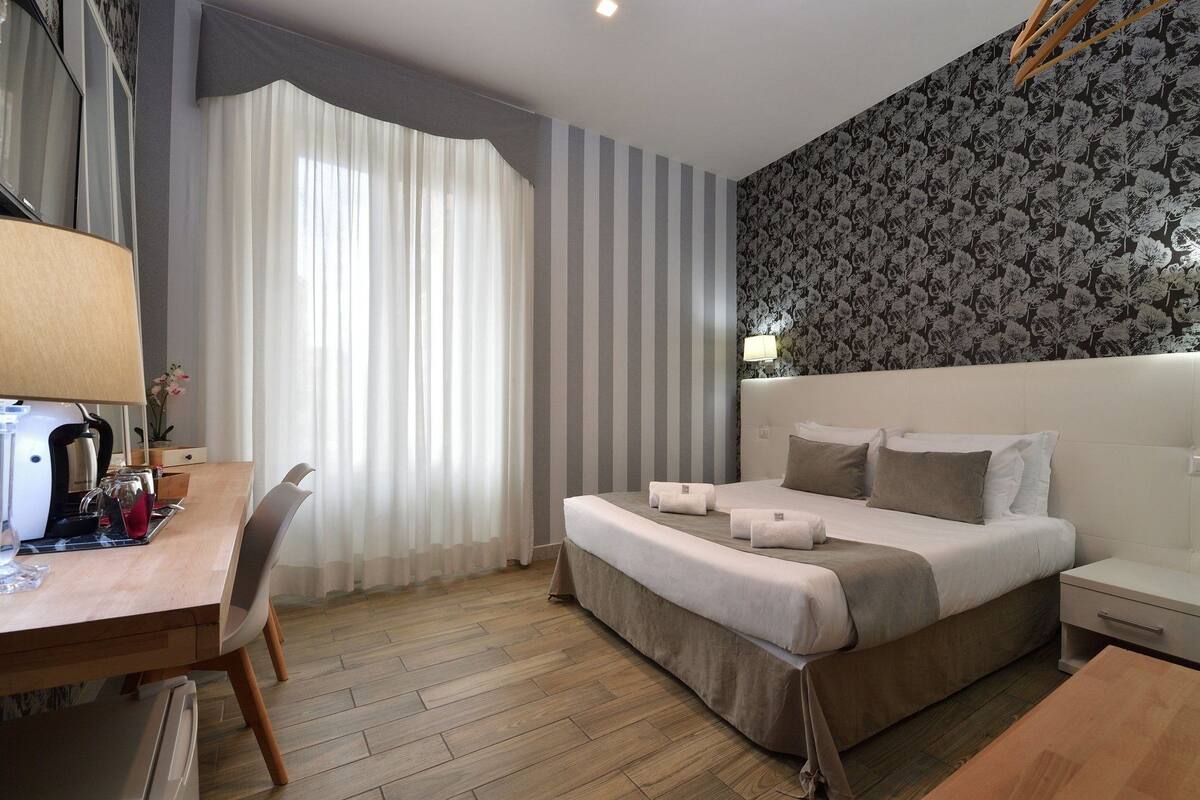 Quarto de hotel com cama, mesa de cabeceira, abajur e escrivaninha com cadeira.