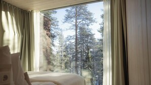 A bedroom with a bed, a window with curtains, and a view of a forest. - Arctic TreeHouse Hotel (Rovaniemi)