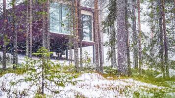 Eine moderne Hütte im Wald mit Glasfassade, umgeben von hohen Bäumen und Schnee.