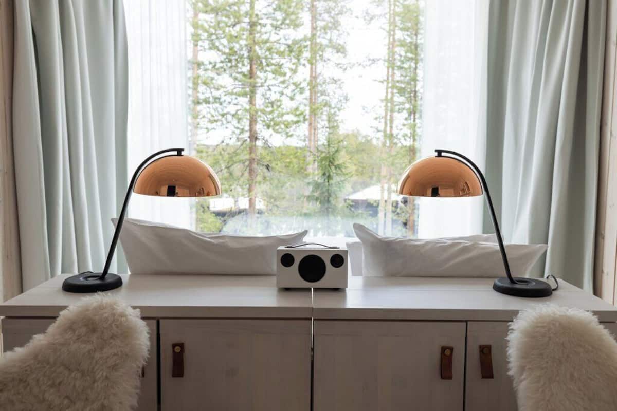 A room with a desk, two lamps, a radio, and a view of trees through the window.