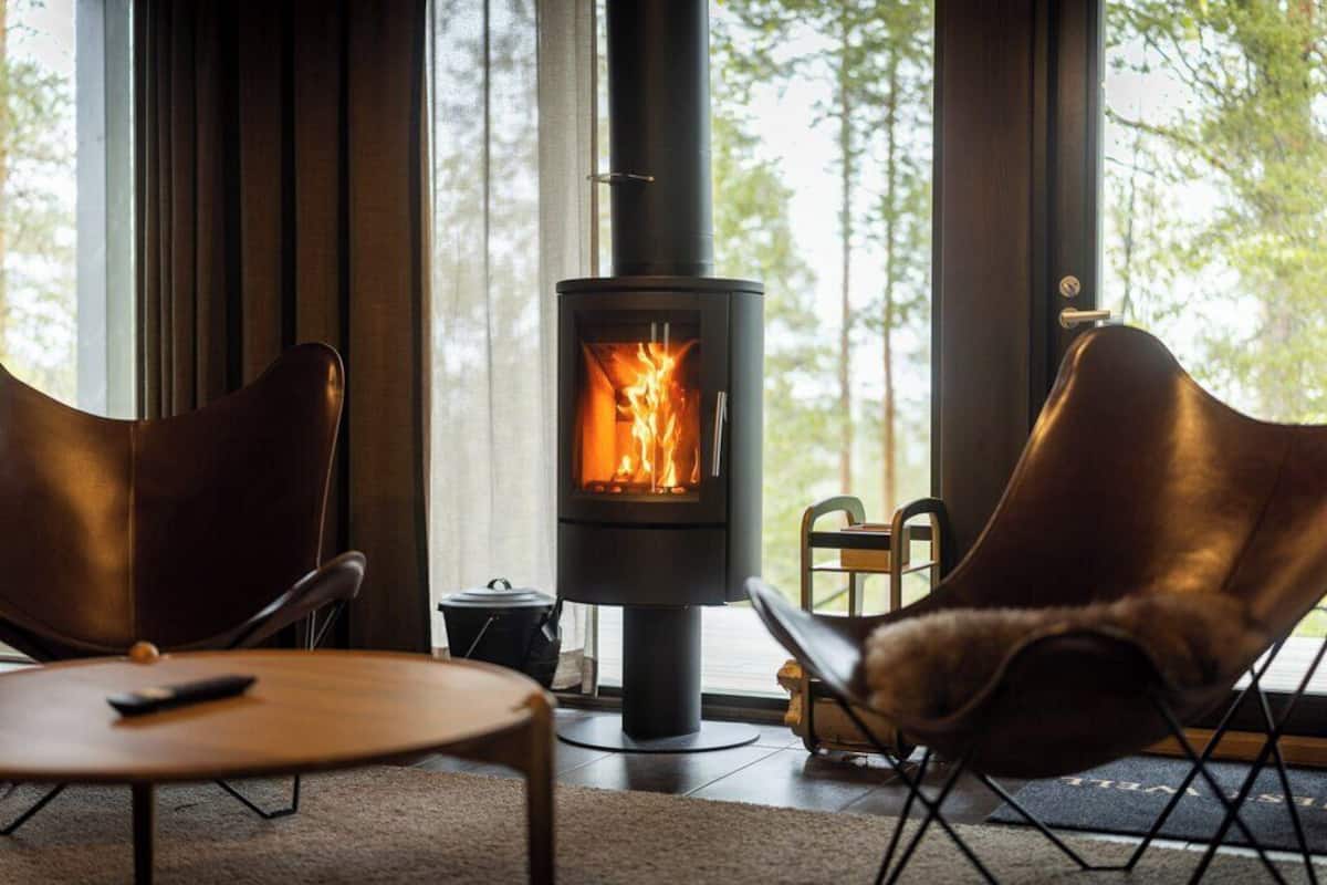 A modern living room with a wood-burning stove, leather chairs, a small table, and a view of trees through large windows.
