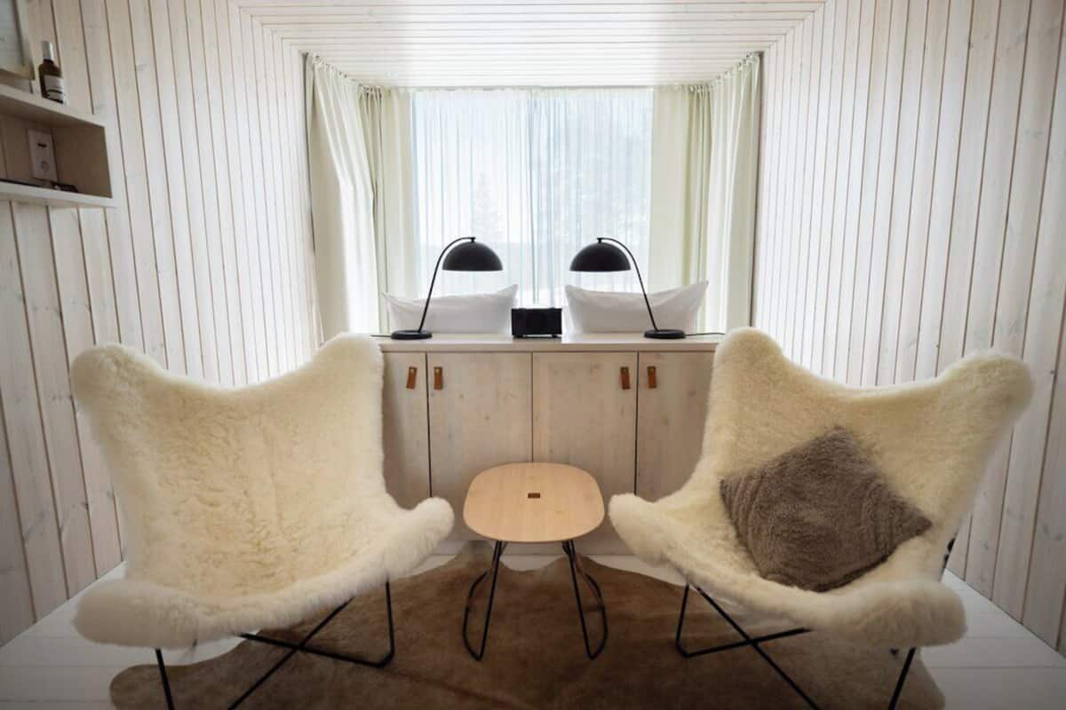 A room with two white furry chairs, a small wooden table, and a radio. There are curtains on the window, and a shelf with bottles on the left.
