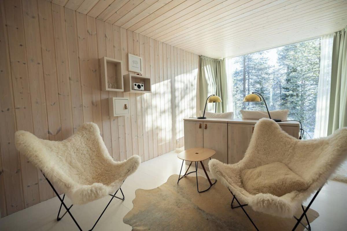 A cozy room with wooden walls, two fluffy chairs, a small round table, a lamp, and a view of trees through a window.