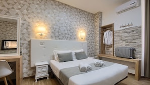 A modern hotel room with a geometric-patterned wall, a large bed with grey bedding, a bedside table, a small bedside table, a desk with a chair, a mirror, a wall-mounted air conditioner, and a coat rack with a jacket. - Domus Pietro Inn (Rome)