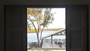 A view through a window with shutters, revealing a lakeside deck with a wooden pergola, outdoor seating, and a tree.