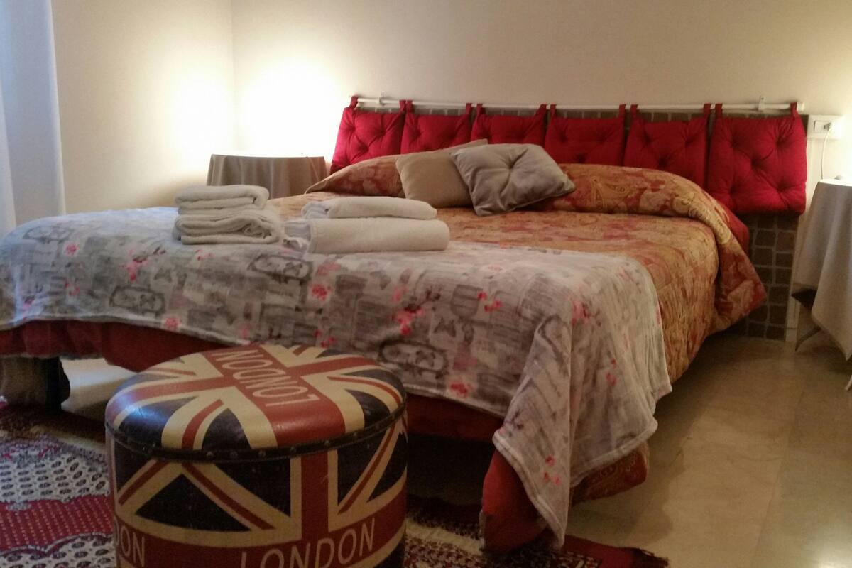 A neatly made bed with a red headboard, a bedside table with a lamp, and a round ottoman with 'LONDON' on it.