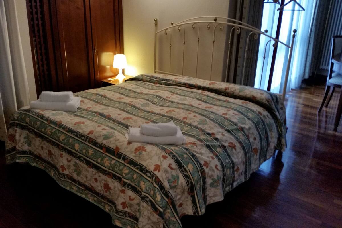 A neatly made bed with floral bedding and two towels, a bedside lamp, and a metal headboard in a room with wooden flooring and a window with curtains.