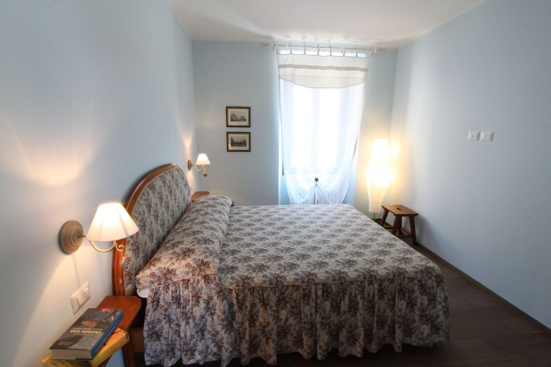Une chambre à coucher avec un lit, deux lampes fixées au mur, une table de chevet avec un livre, et une fenêtre avec des rideaux légers.