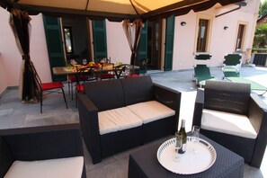 Un patio exterior con mobiliario de mimbre, una mesa de comedor dispuesta con platos, vasos y una botella de vino.