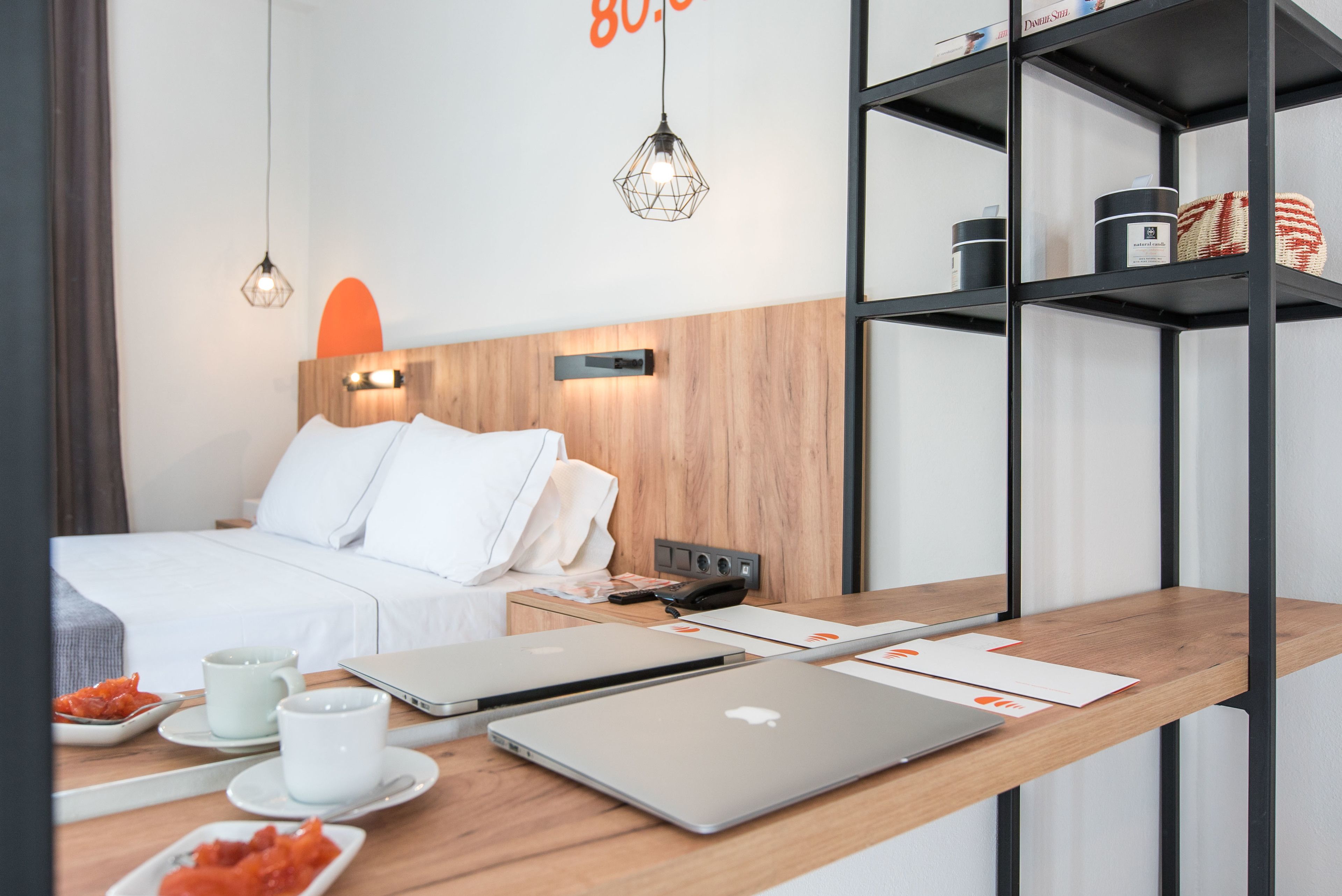 A modern hotel room with a wooden headboard, a bed with white linens, a desk with two laptops, a coffee setup, and a shelf with decorative items.