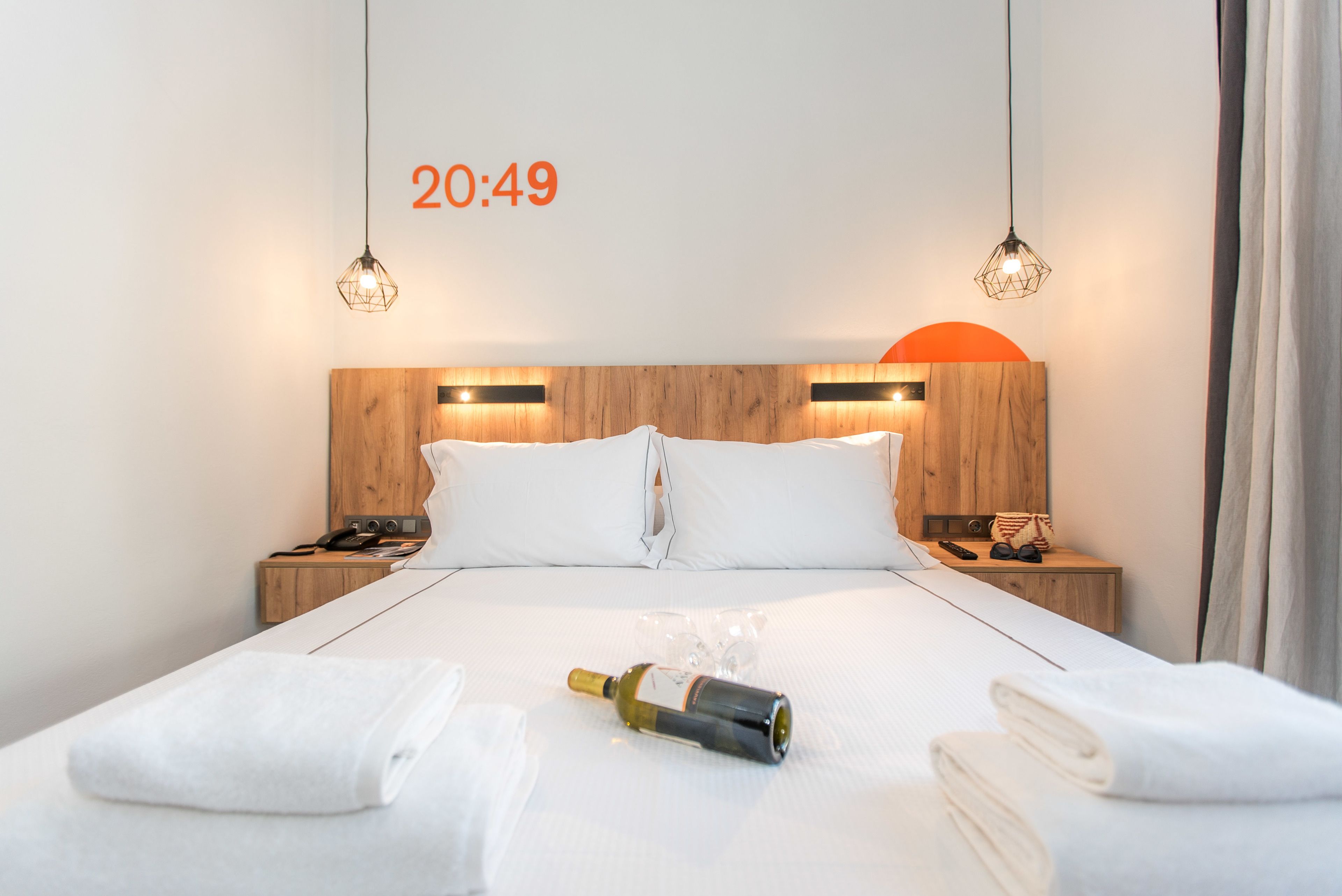A hotel room with a wooden headboard, two bedside lamps, a bed with white linens, a bottle of wine, and a clock displaying 20:49.