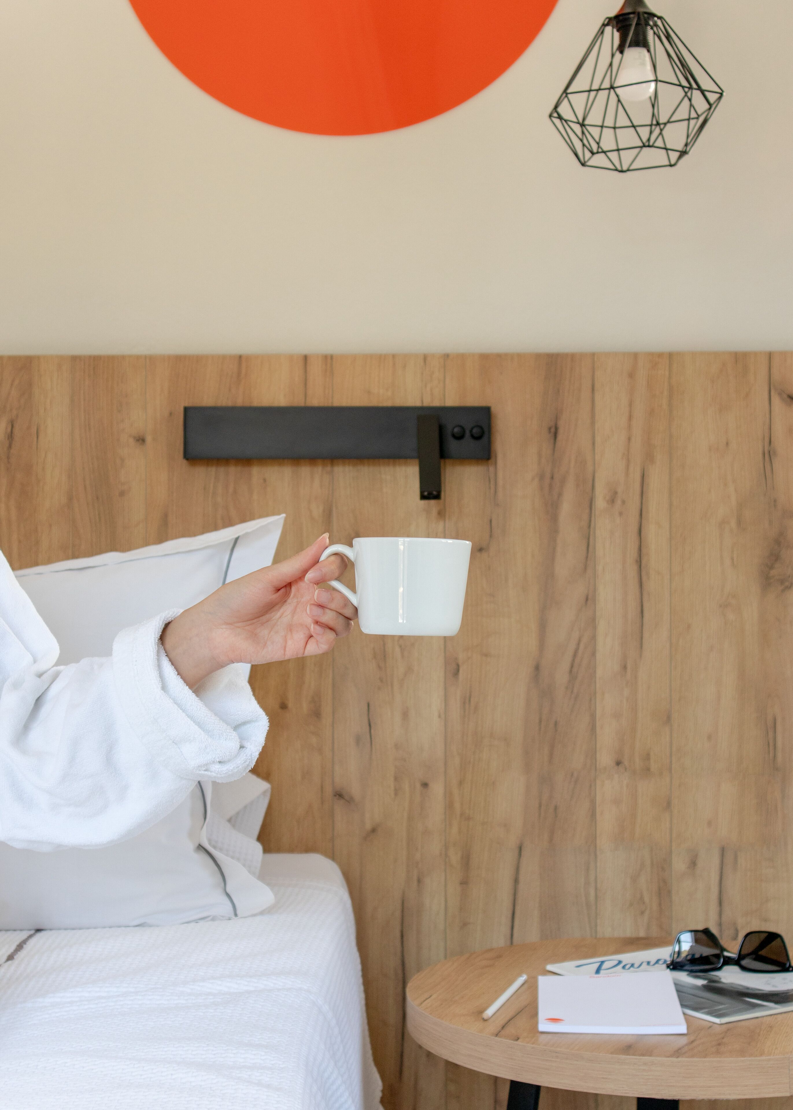 Une personne tenant une tasse blanche, un mur en bois, un lit avec une literie blanche, une petite table ronde avec un livre, des lunettes et un stylo.