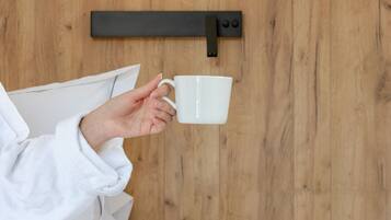 Une personne tenant une tasse blanche, un mur en bois, un lit avec une literie blanche, une petite table ronde avec un livre, des lunettes et un stylo.