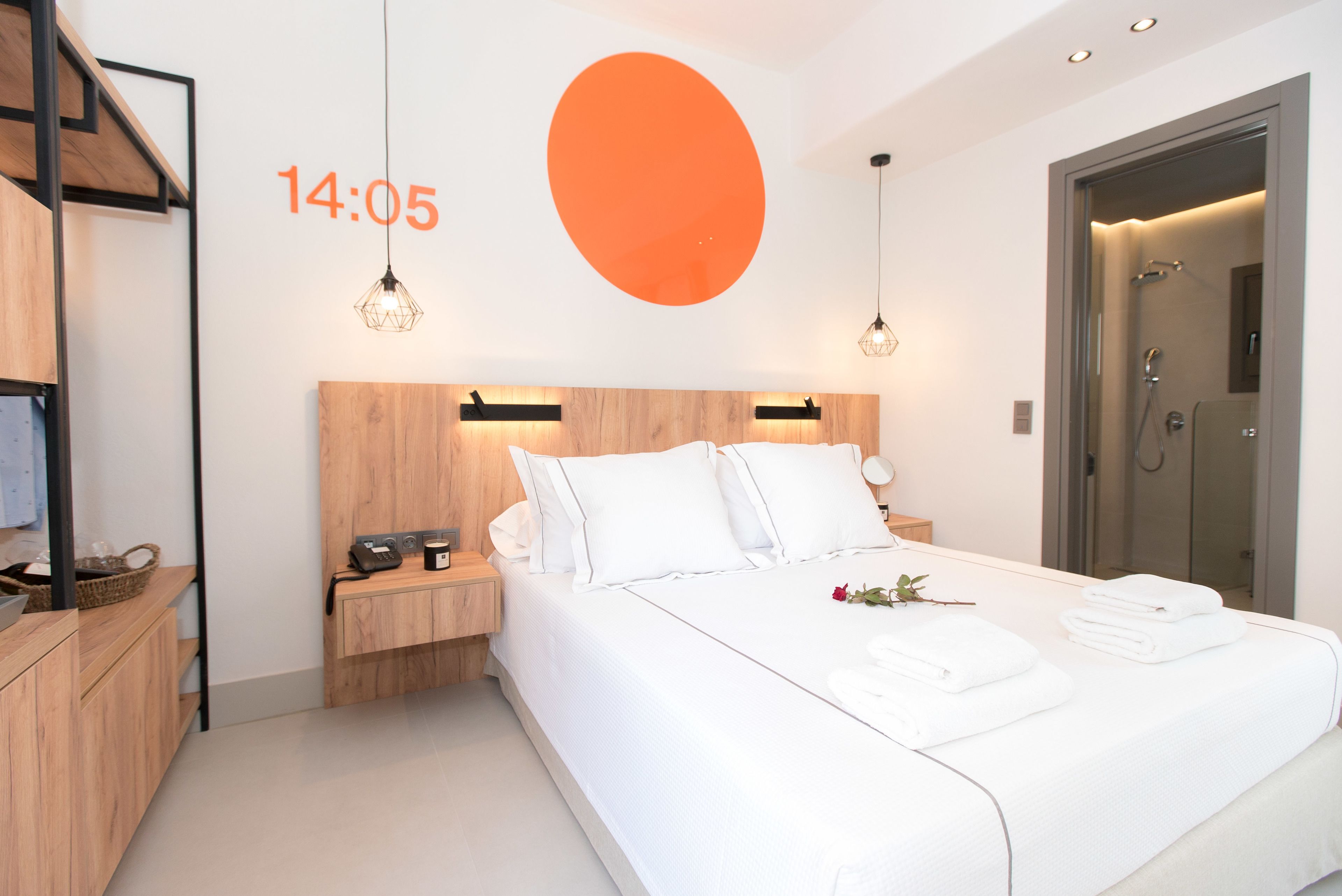 Una habitación de hotel moderna con cabecera de madera, una cama grande con ropa blanca, una mesita de noche con lámpara y un reloj digital que marca las 14:05.