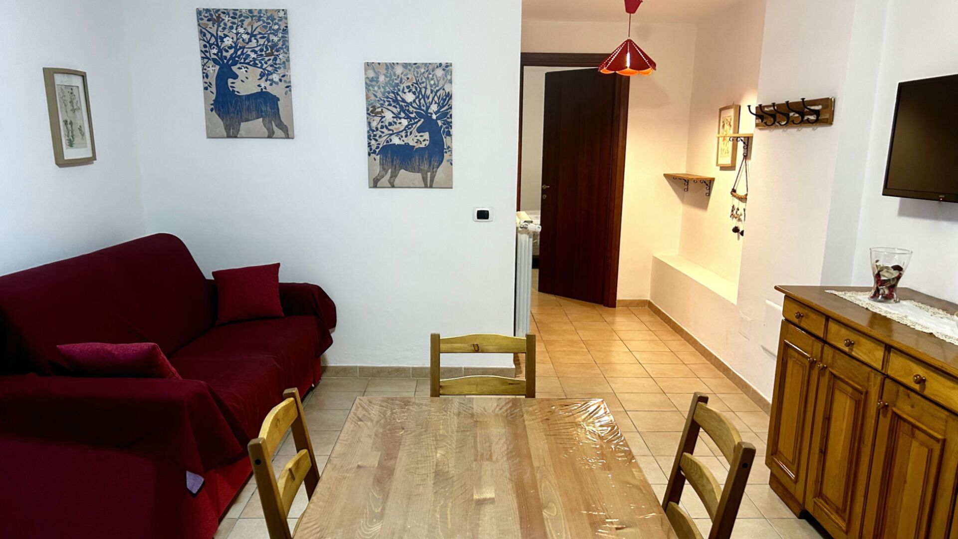 A living room with a red sofa, wooden dining table, and two wooden chairs. There are two blue paintings on the wall and a television on the right.
