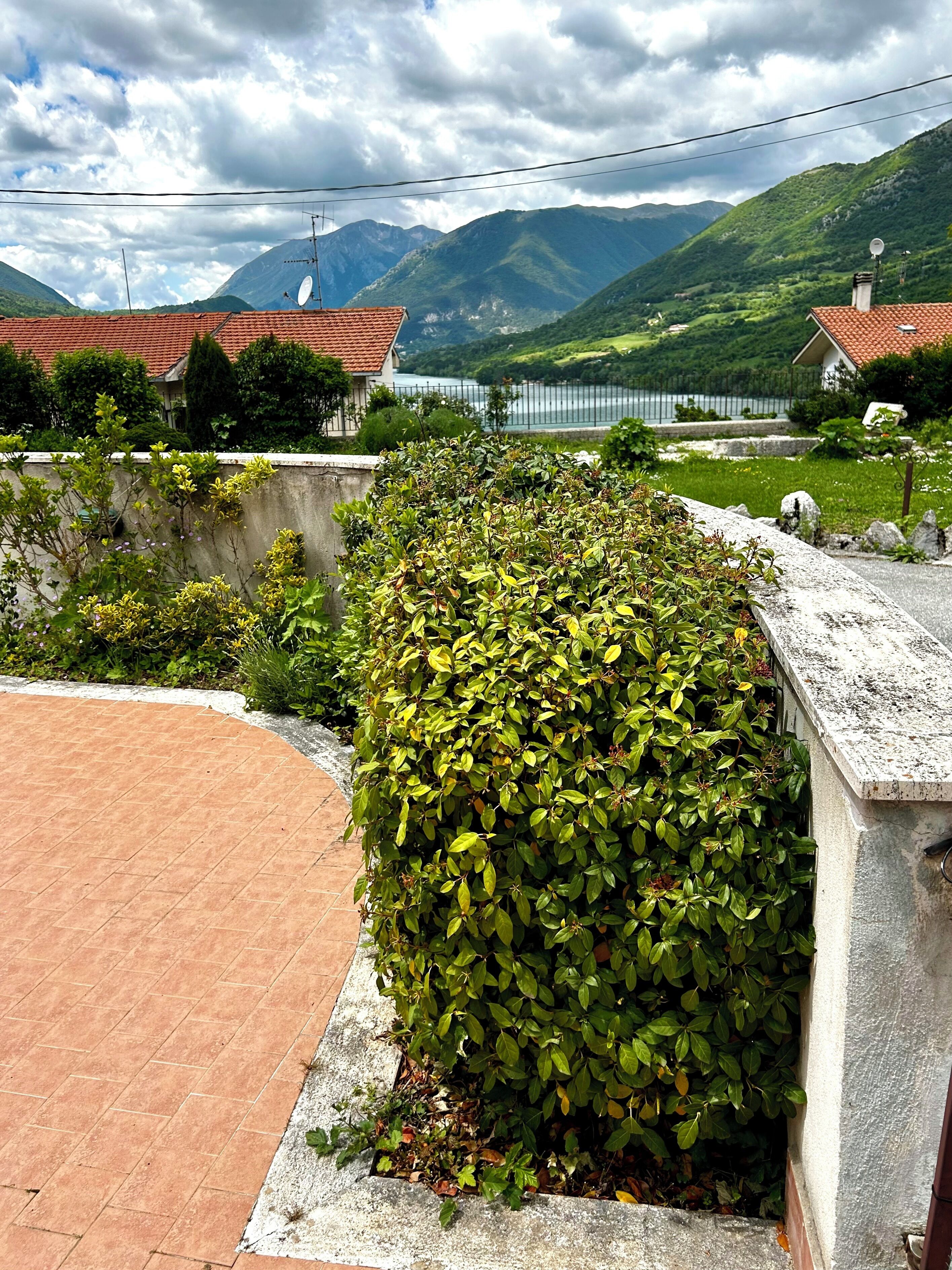 Pátio pavimentado com sebe, muros de pedra e vista para montanhas e um lago.