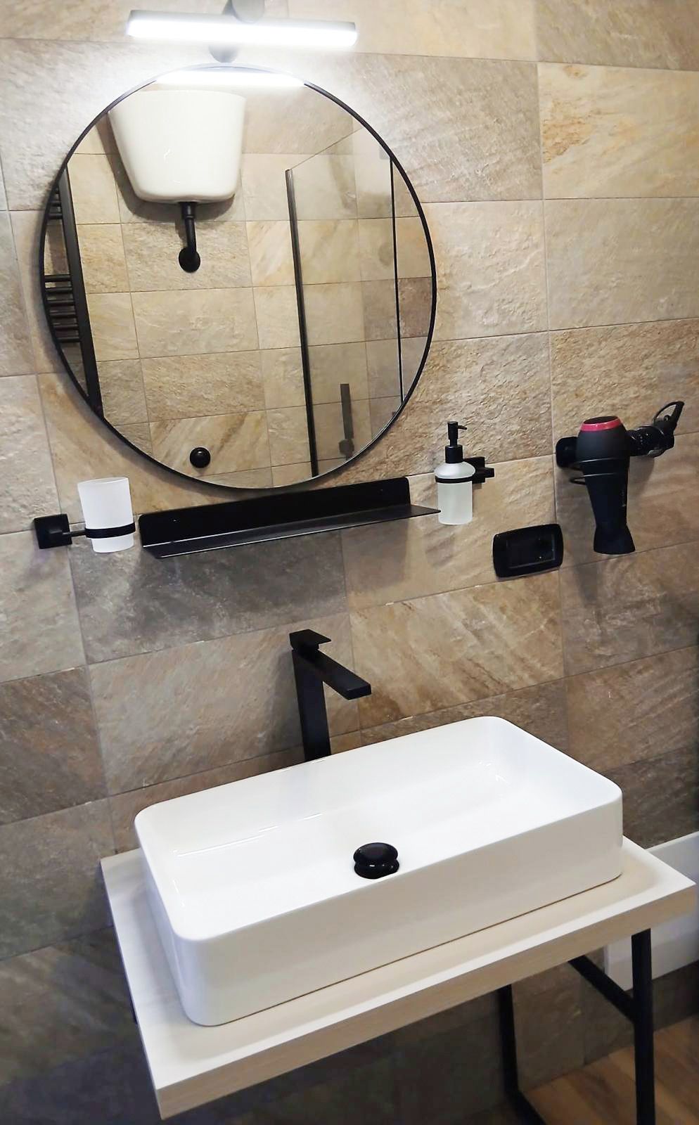 A bathroom with a white sink, a round mirror, a handheld showerhead, and a soap dispenser.