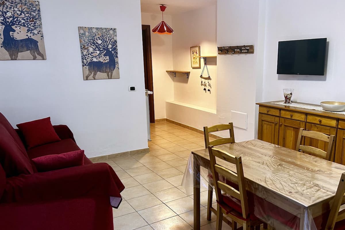 Un salon avec un canapé rouge, une table à manger en bois avec des chaises, un coin cuisine avec des meubles en bois, un téléviseur fixé au mur et des œuvres d’art accrochées aux murs.