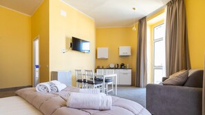 A hotel room with a large bed, a sofa, a dining area with chairs, a TV mounted on the wall, and a kitchen area. - B&B Trianon (Naples)