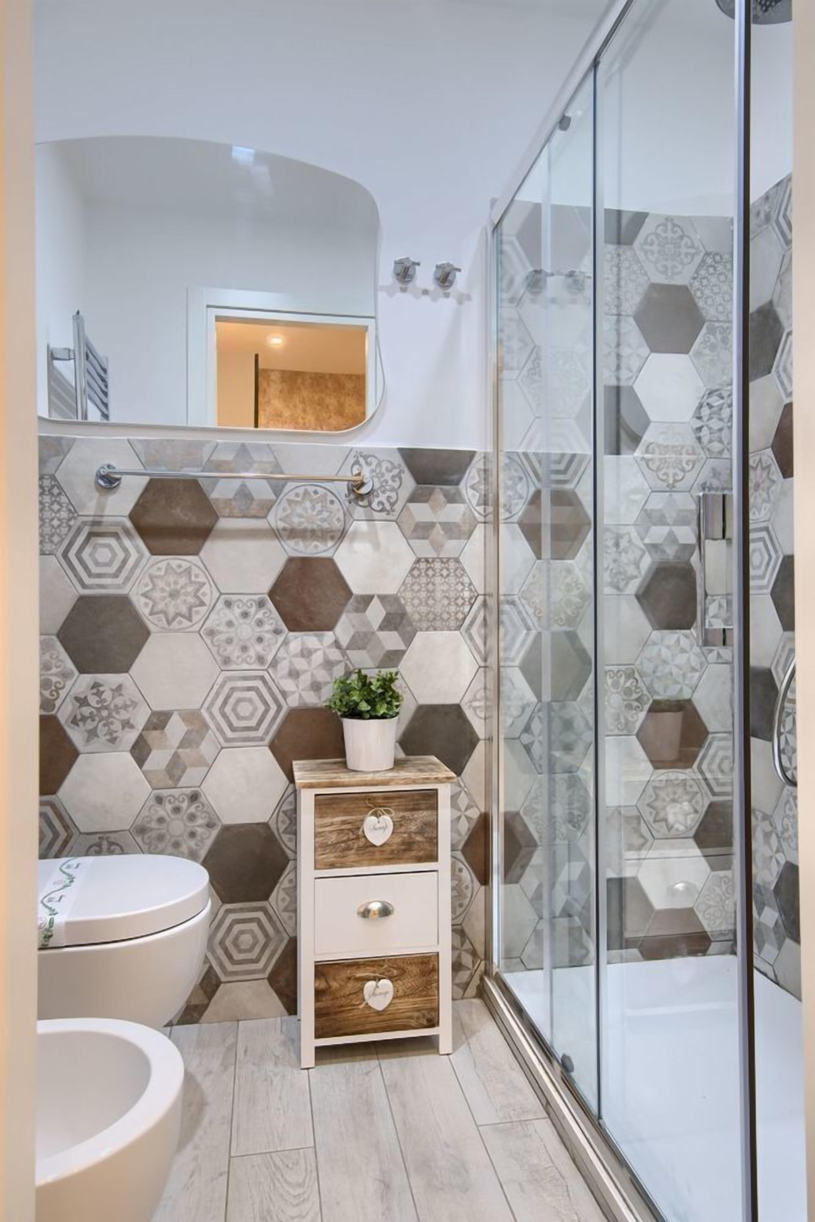 A bathroom with a hexagonal tile backsplash, a wooden vanity with drawers, a toilet, and a glass shower enclosure.