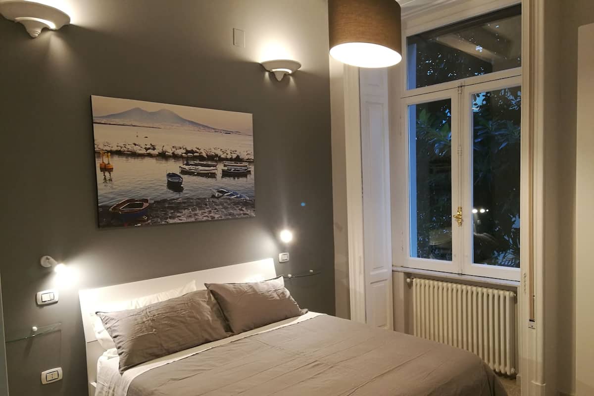 Un dormitorio con cama, lámparas de noche, ventana con vista a árboles y un cuadro en la pared.