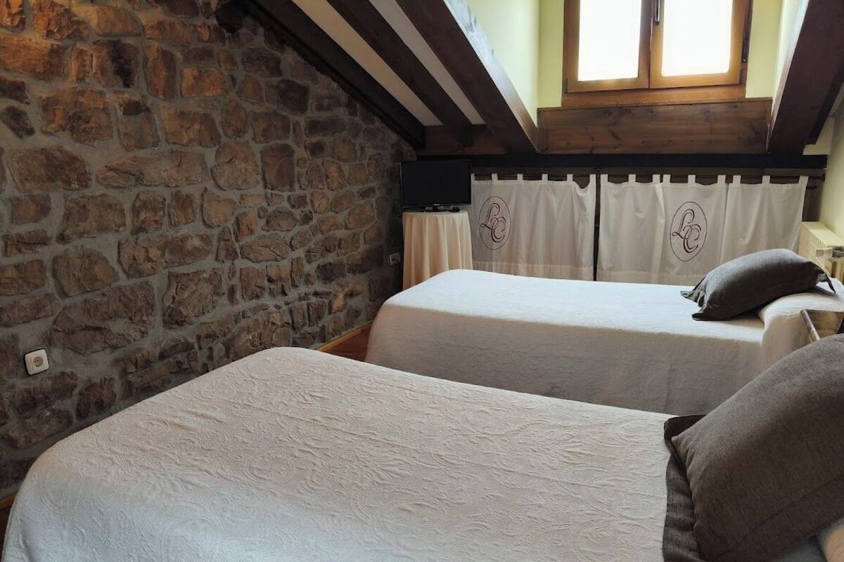 Une chambre avec deux lits, des murs en pierre, un plafond en bois et une fenêtre avec des rideaux.