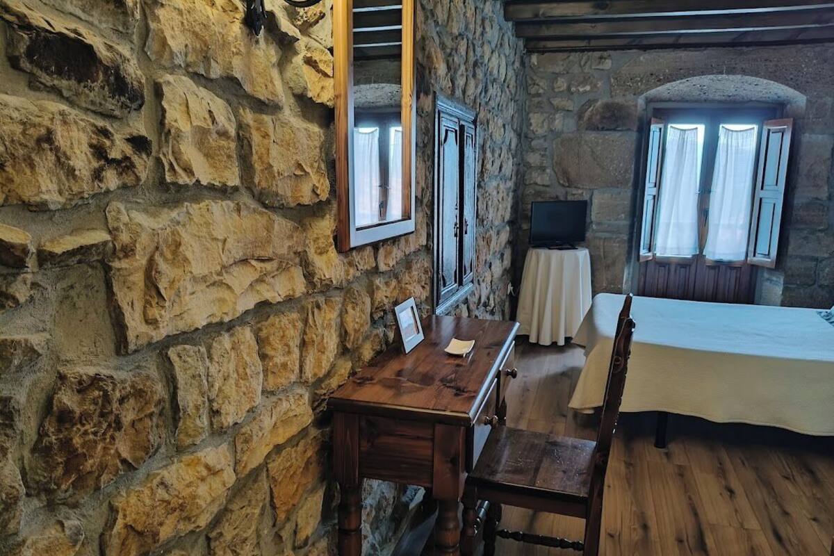 Une pièce aux murs de pierre, avec un bureau en bois, une chaise, un lit recouvert d’une couverture blanche et un téléviseur posé sur une petite table.