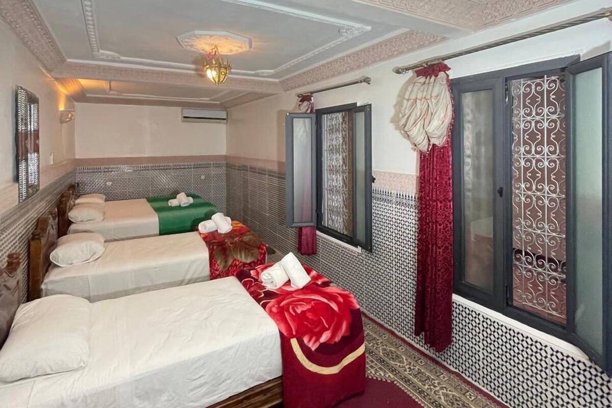 A hotel room with four beds, a large window with decorative curtains, and a patterned carpet.