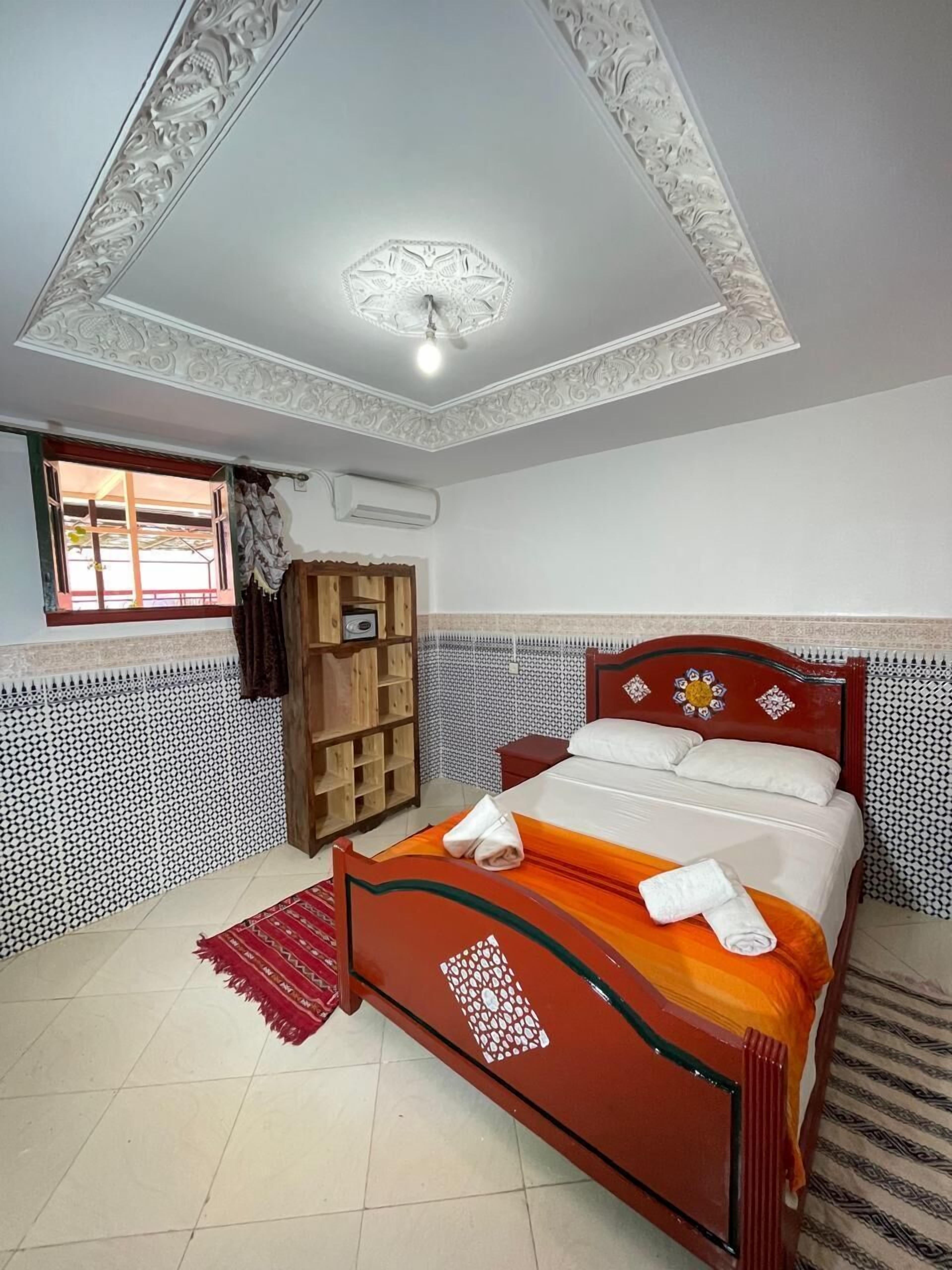 Un dormitorio con cama de madera, cabecera estampada en rojo y blanco, techo decorativo y ventana con cortinas.