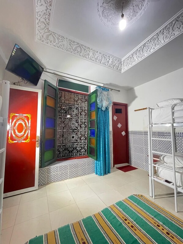 Una habitación con una cama litera, un televisor montado en la pared, un ventanal decorativo y un techo ornamentado.