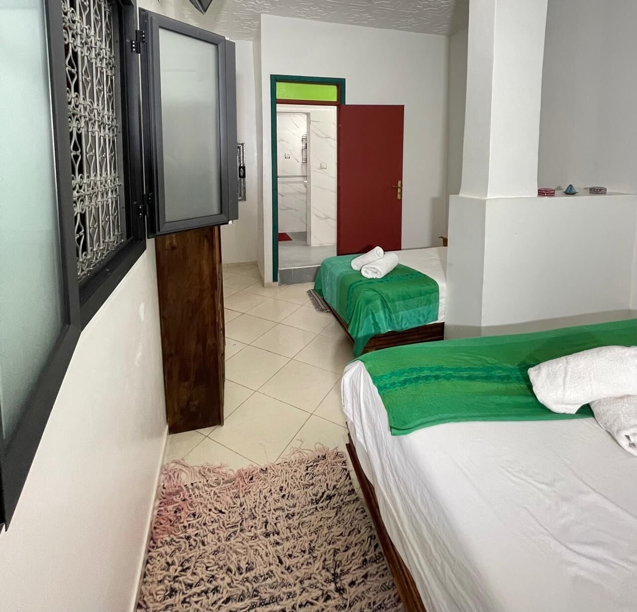 A hotel room with two single beds, a wooden wardrobe, a mirror, and a view of a bathroom with a red door.