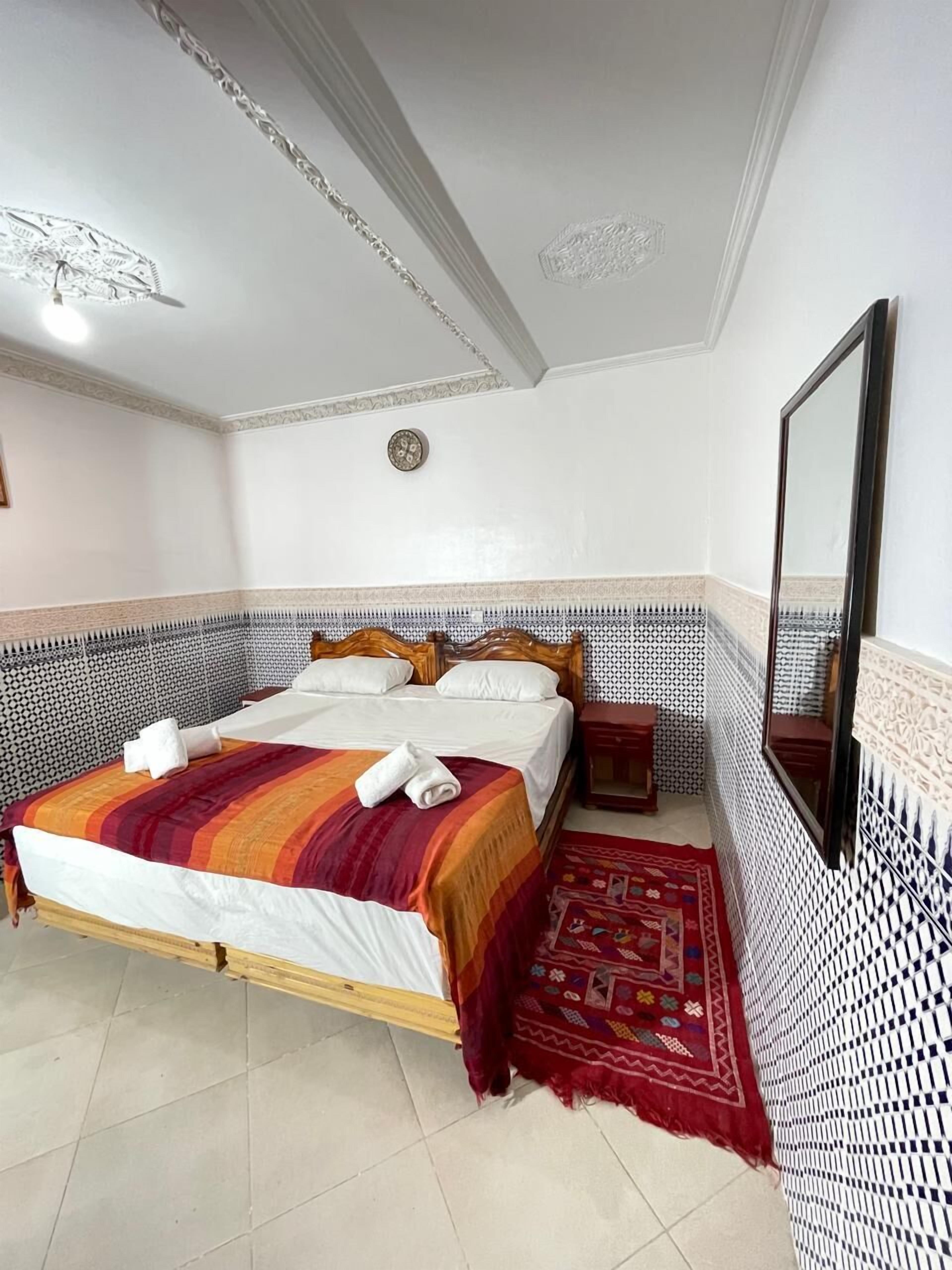 A neatly made bed with a striped blanket and towels, a wooden headboard, a small bedside table, a mirror, and patterned tile walls.