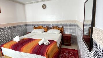 A neatly made bed with a striped blanket and towels, a wooden headboard, a small bedside table, a mirror, and patterned tile walls.