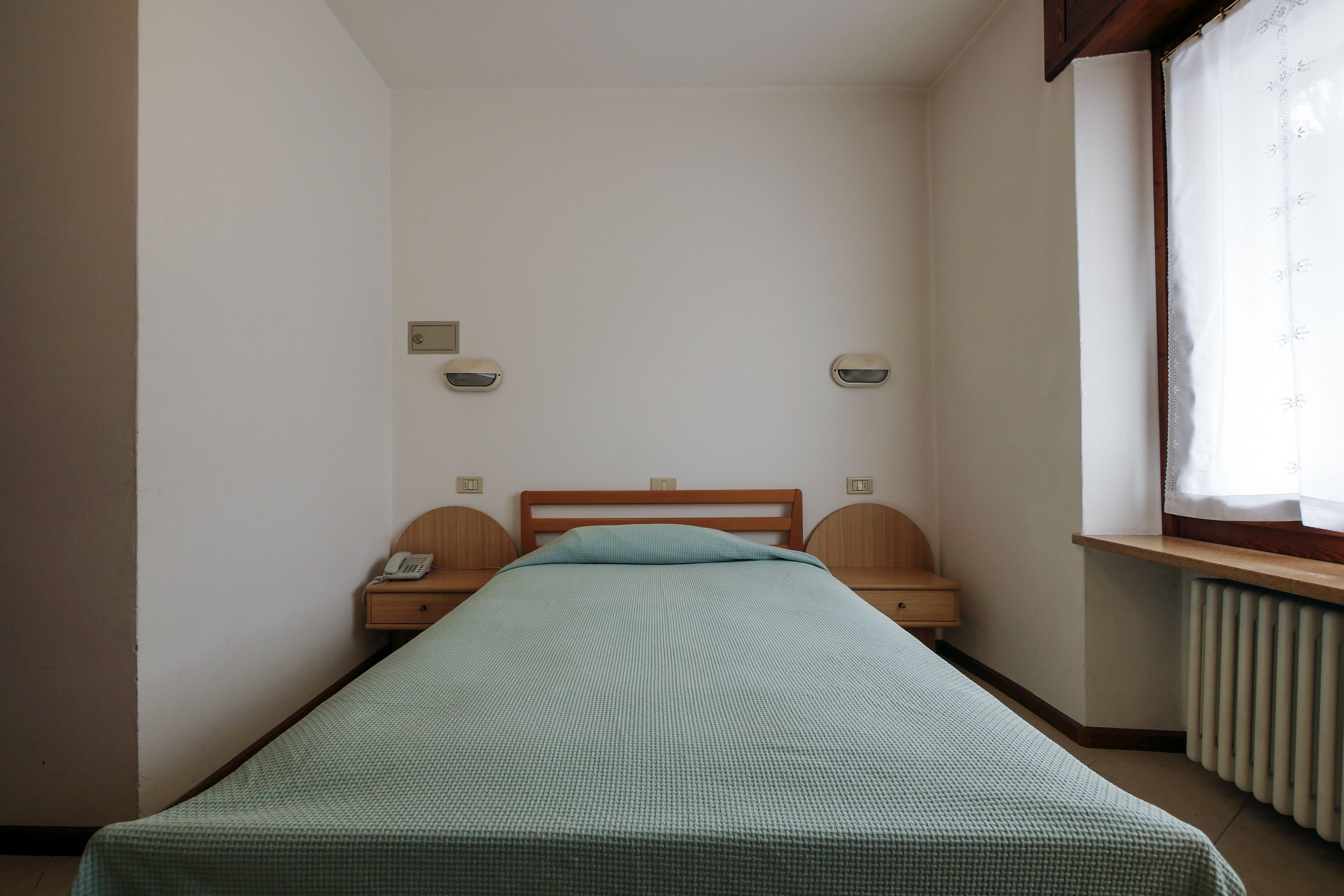 A small, single-bed room with a wooden headboard, bedside tables, and a window with a curtain.