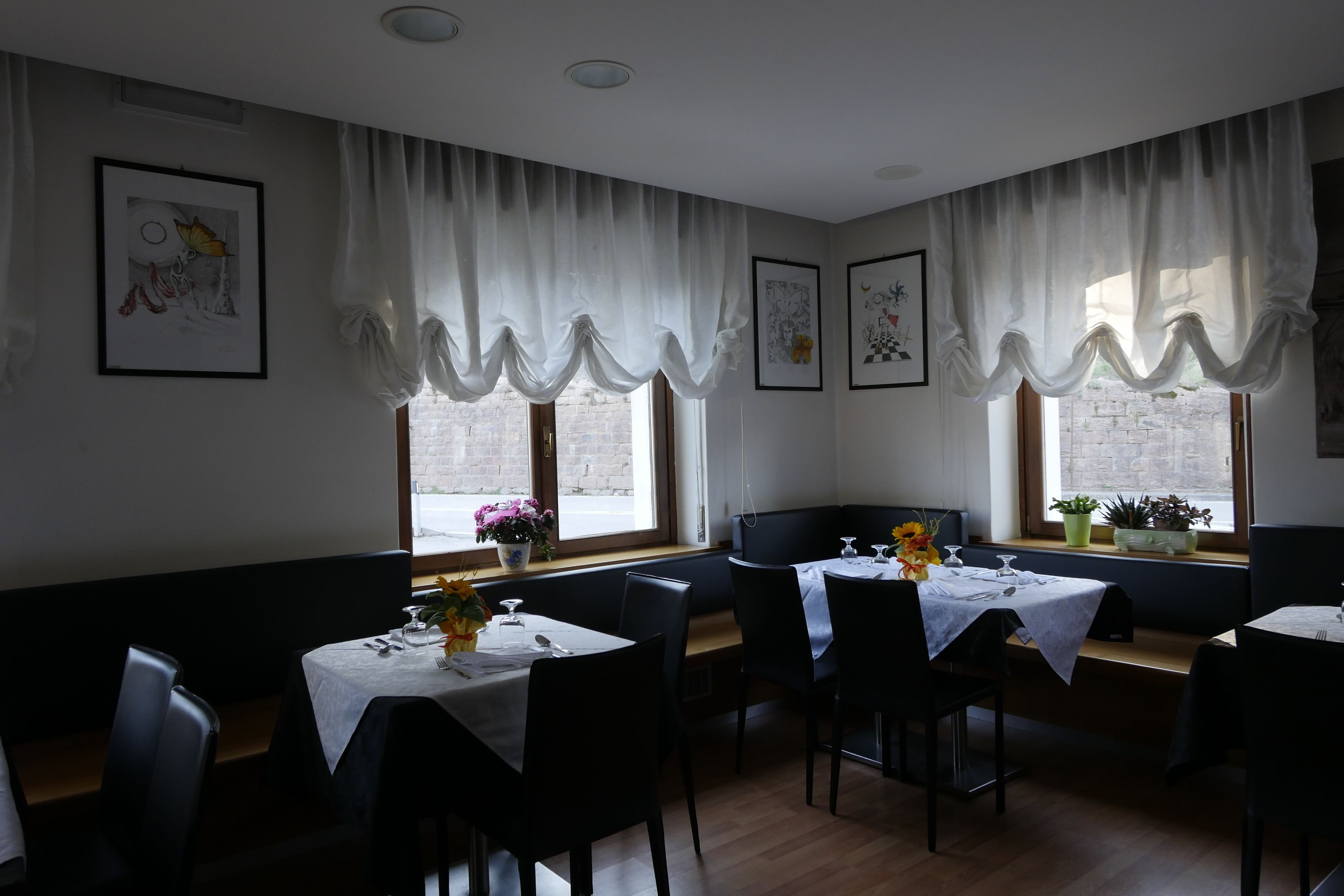 A dining area with tables set for two, white curtains, and artwork on the walls.