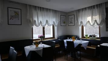 A dining area with tables set for two, white curtains, and artwork on the walls.