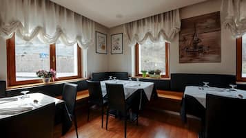 Un restaurante con suelo de madera, sillas negras, manteles blancos y amplios ventanales con cortinas.