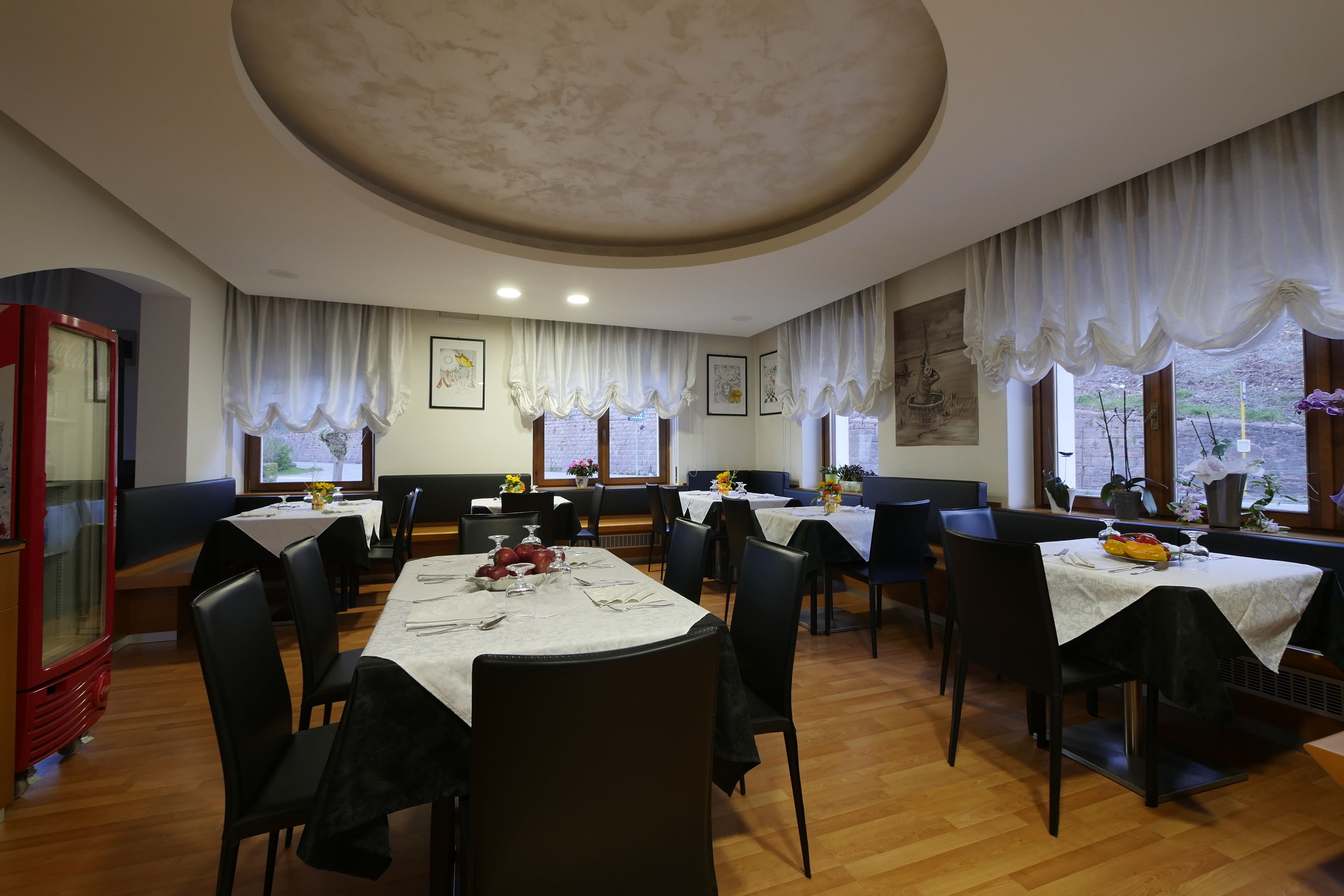 Un restaurant avec des tables rondes, des chaises noires et des nappes blanches. Il y a des fenêtres avec des rideaux et des œuvres d’art accrochées aux murs.