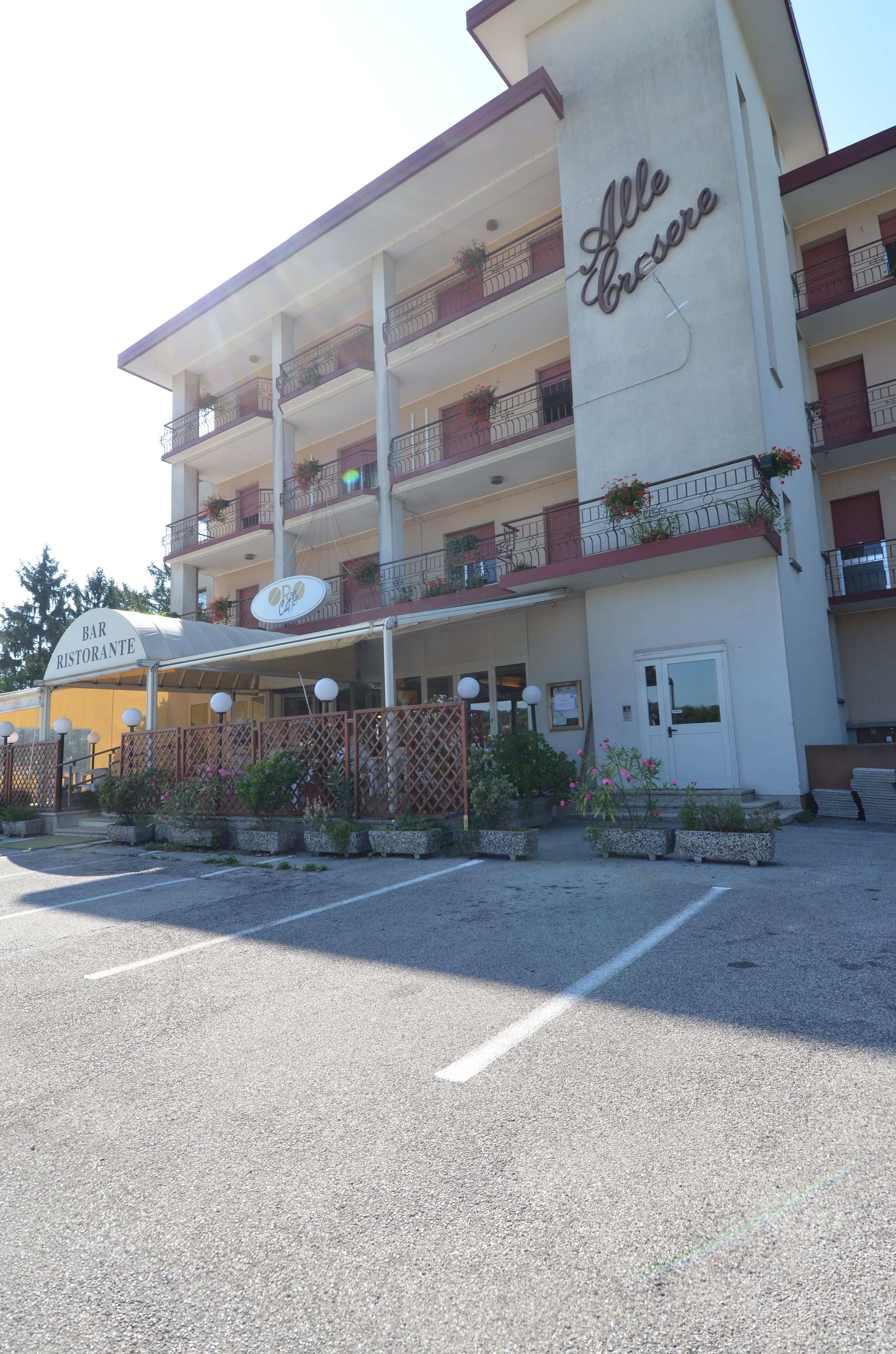 A multi-story building with a sign reading "Bar Ristorante" and "Alle Crocere." It features balconies with plants and a parking area in front.