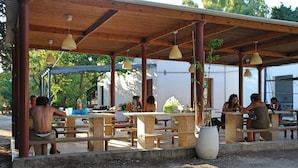 An outdoor café with wooden tables and benches, covered by a wooden roof with hanging pendant lights. - Krios Beach Camping (Paros)