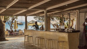 An outdoor bar area with a stone floor, wooden stools, and umbrellas. - Krios Beach Camping (Paros)