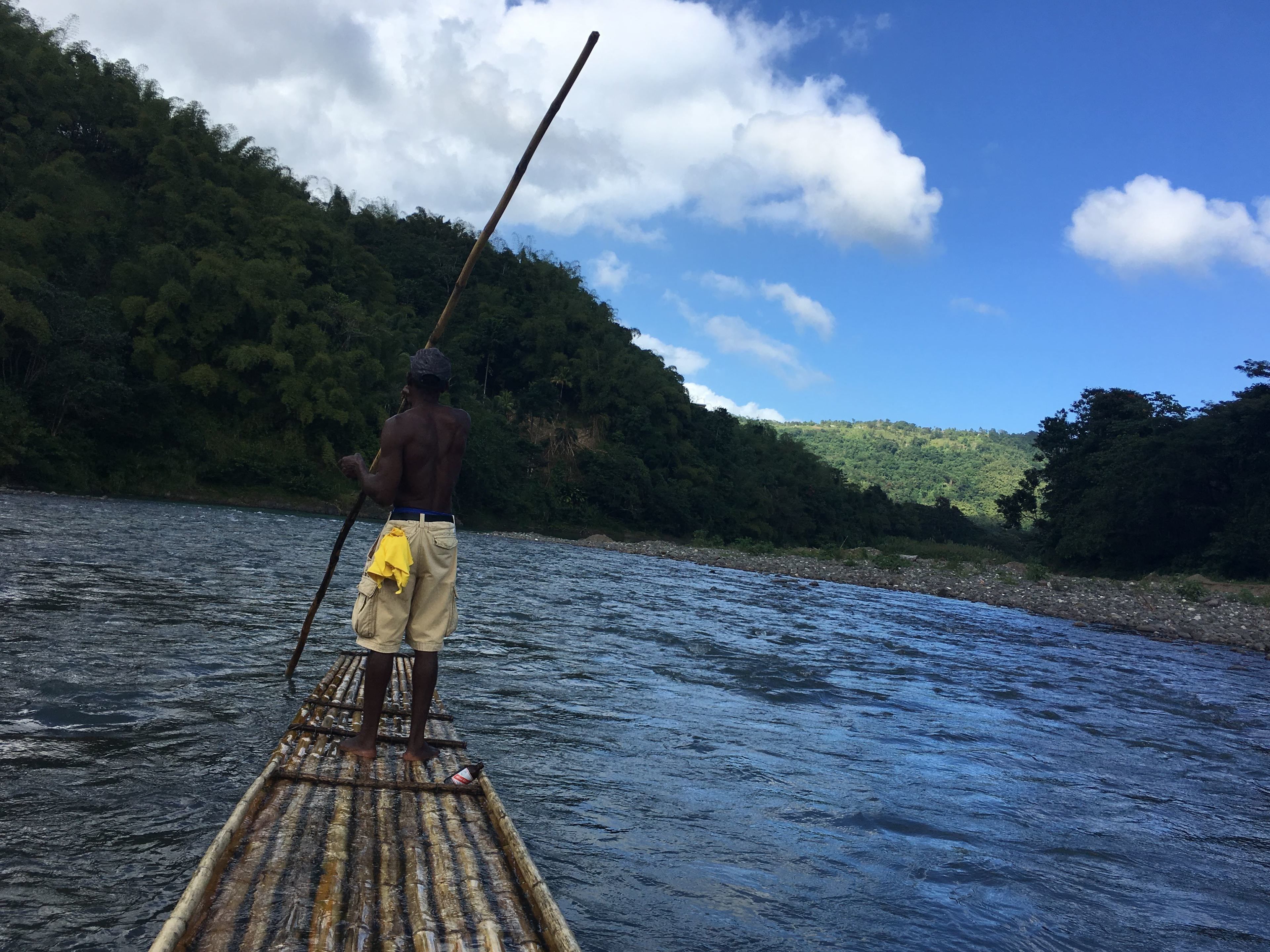 Une personne se tient sur un radeau de bambou dans une rivière, entourée d’une végétation luxuriante et sous un ciel dégagé.