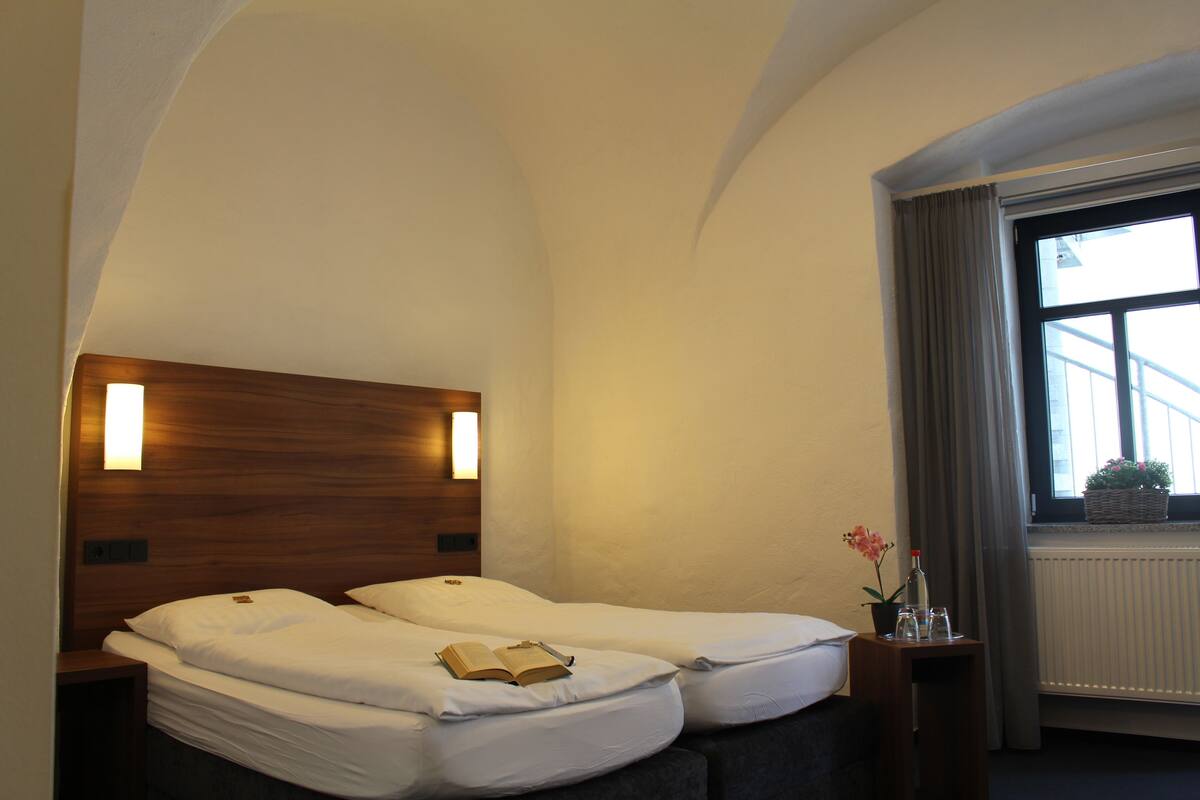 A hotel room with two beds, a wooden headboard, a nightstand with a lamp, a window with curtains, and a potted plant on the windowsill.