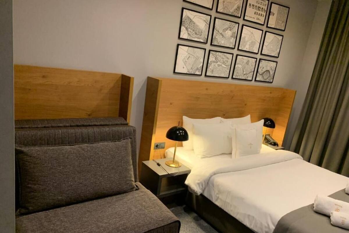 A hotel room with a bed, a nightstand, a lamp, a chair, and a wall with framed pictures.