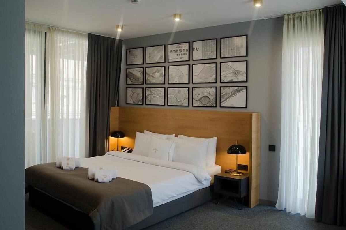 A modern hotel room with a large bed, two bedside tables with lamps, a headboard with artwork, and sheer curtains.