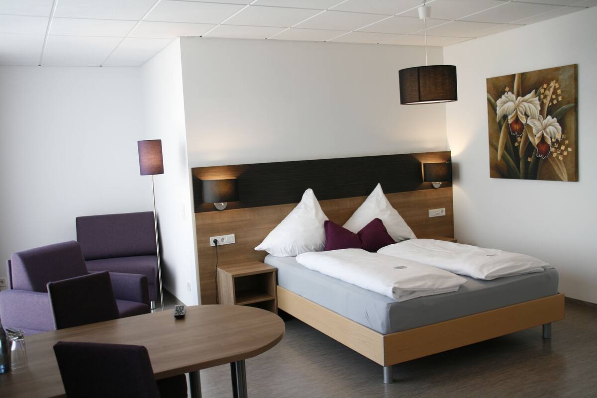 Habitación de hotel moderna con una cama de madera, una mesita de noche pequeña, una silla y una mesa redonda.