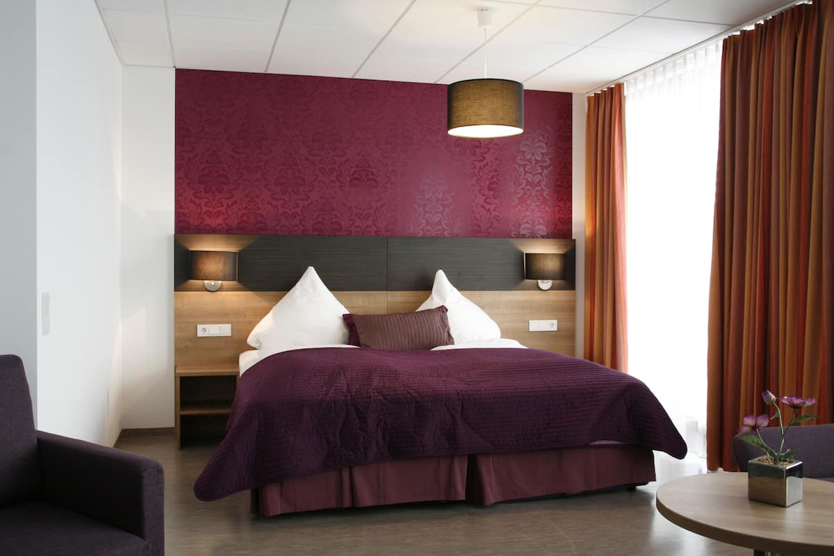 Habitación de hotel moderna con una cama grande, cabecera de madera, mesita de noche, una silla y una mesa redonda con un florero.