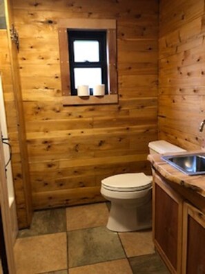 A small, wooden-paneled bathroom with a toilet, a window with two towels, and a sink.