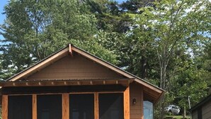 A wooden cabin with a deck, a grill, and a vehicle parked nearby. - Riverbay Adventure Inn (Chippewa Bay)